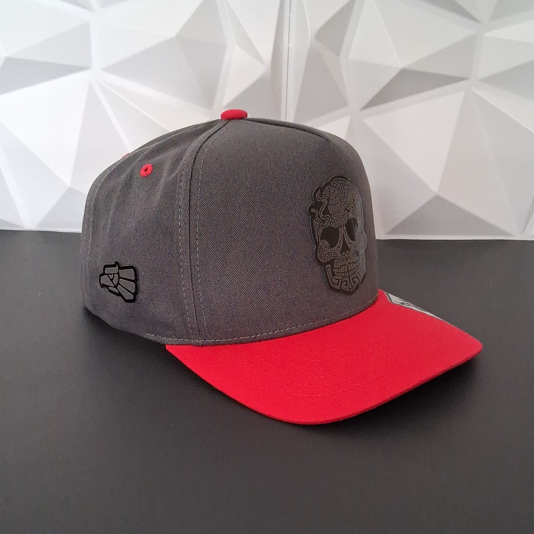 Gray and red baseball cap with a black logo on a dark surface and geometric patterned wall background